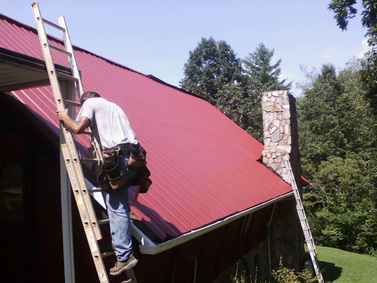 Roofer on a ladder beside a red metal roof and stone chimney — Soffit & Fascia Repair by Roofing Brothers in Tulsa